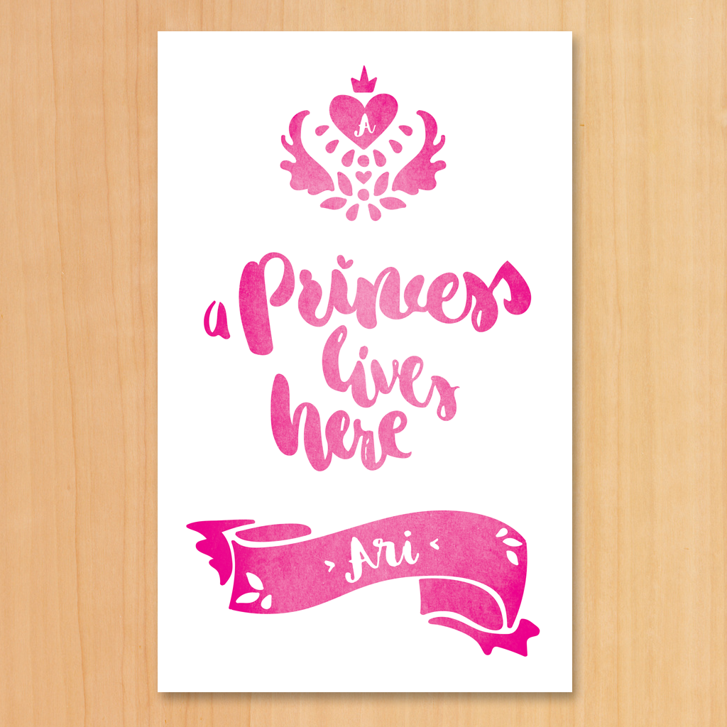 A white and pink print with a royal crest and text "A Princess Lives Here". Set your girl's initial in the heart inside the crest and her name in the decorative banner below.